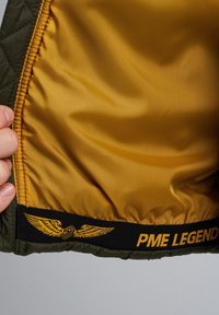 Grüne gesteppte Jackeninnenseite mit glatter gelber Fütterung, ausgestattet mit einem schwarzen Band, das mit "PME LEGEND" und einem geflügelten Logo verziert ist.