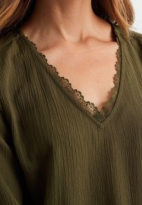 Olivgrön blus med en texturerad tygkvalitet. Har en V-ringning med delikat spetskant och svaga vågmönster.