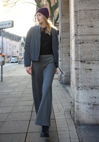 Modella che indossa una giacca bomber imbottita grigia, top nero e pantaloni grigi a gamba larga, con un berretto bordeaux e stivaletti neri, che cammina su un marciapiede.