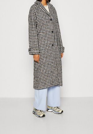 Femme portant un long manteau pied-de-poule noir, blanc et beige sur un pantalon large bleu clair et des chaussures de sport beige.