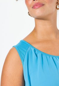 En närbild av en blå, ärmlös topp med mjukt tyg och veck längs halsringningen samt en slät, rund axeldesign.