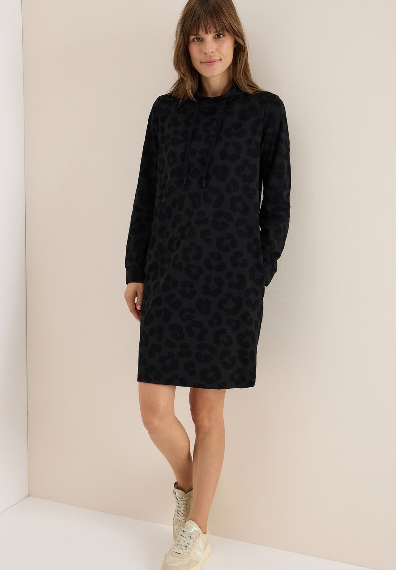 Svart hoodieklänning med en texturerad leopardtryck, med långa ärmar, avslappnad passform och sidofickor, upp tillsammans med ljusa sneakers.