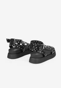 Black sandals with studded straps and a flat sole. Features a wide ankle strap and buckle closure. Smooth texture and chunky design.