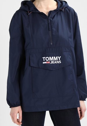 Granatowa kurtka z kapturem, zapinana na zamek z przodu, z dużą kieszenią z logo "Tommy Jeans" w kolorze białym, noszona przez osobę.