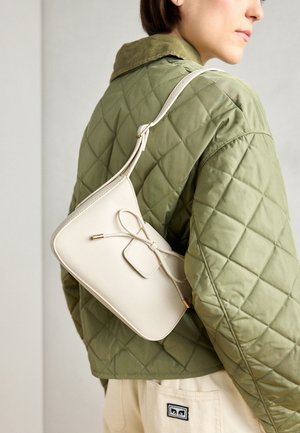 Bolso cruzado de piel color beige con detalle de lazo, herrajes dorados y una sola correa ajustable, llevado sobre una chaqueta de lana acolchada verde.