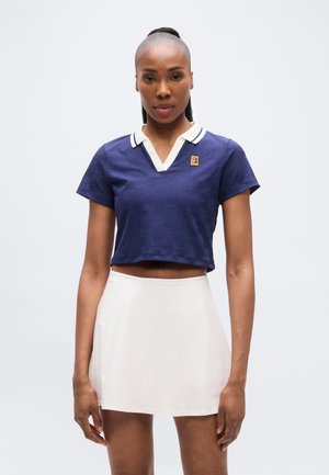 Top court polo bleu marine avec un motif texturé, col blanc et écusson logo ; associé à une jupe patineuse crème. Tissu lisse et manches courtes.