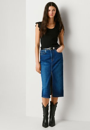 Kvinne med langt mørkt hår iført svart ermeløs topp, blå denim midiskjørt med frontsplitt, svart belte og svarte støvler som når midt på leggen, stående.