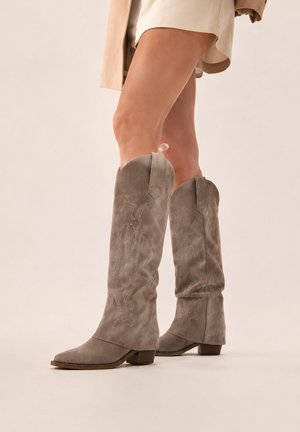 Jambes portant des bottes en daim beige montant jusqu’au genou avec des motifs brodés subtils et des talons blocs, associées à une jupe légère.