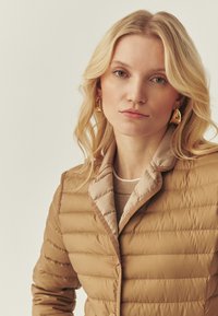 Veste matelassée beige avec un col à revers couleur châtain clair. Texture douce, matelassage horizontal, fermeture à boutons et doublure intérieure lisse.