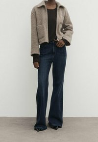 Kvinna som bär en beige fluffig jacka över en mörk topp, kombinerad med högmidjade mörka utsvängda jeans och spetsiga svarta skor.