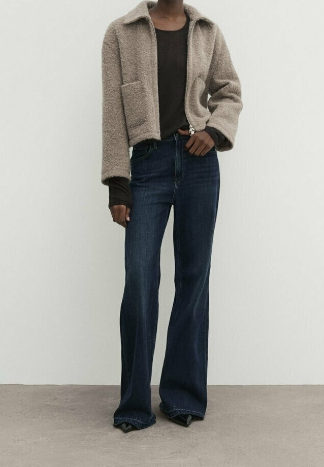 Kvinna som bär en beige fluffig jacka över en mörk topp, kombinerad med högmidjade mörka utsvängda jeans och spetsiga svarta skor.