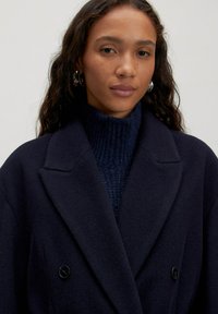 Marineblauwe dubbelzijdige jas met grote revers, gemaakt van een zacht materiaal, gedragen over een donker ribbel turtleneck trui. Zilveren ringoorbellen zichtbaar.