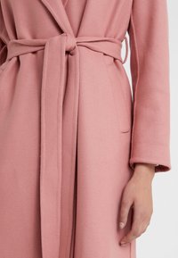 Růžový vlněný kabát s texturovaným povrchem, který má zavinovací design, dlouhé rukávy a pásek uvázaný v pase. Obsahuje boční kapsy.