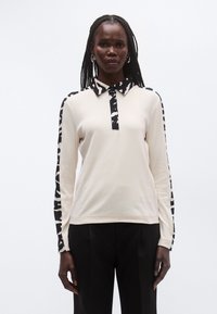 Krämfärgad långärmad pikétröja med svart- och vitmönstrad krage och ärmar, med knappslå och mjuk textur.