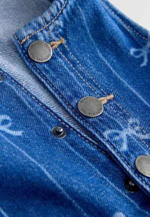 Detailní záběr modrého denimového materiálu s kovovými patentními knoflíky a béžovým prošitím, který ukazuje texturu a detaily knoflíků.
