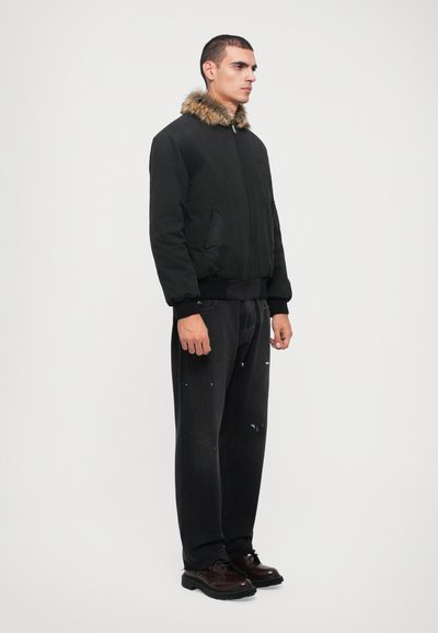 Svart bomberjacka med fake pälskrage, dragkedjeslutning och ribbade muddar, i kombination med slitna svarta jeans och bruna skor.