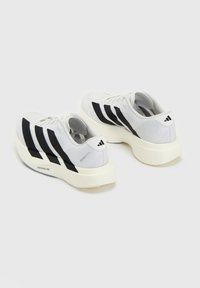 Paire de baskets blanches de sport avec semelles épaisses et design à trois bandes noires vues de l'arrière et de côté sur un fond uni.