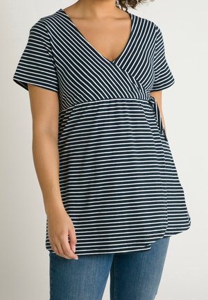 Mujer con una blusa de maternidad cruzada de rayas blancas y negras, de manga corta y jeans azules, de pie frente a un fondo liso.