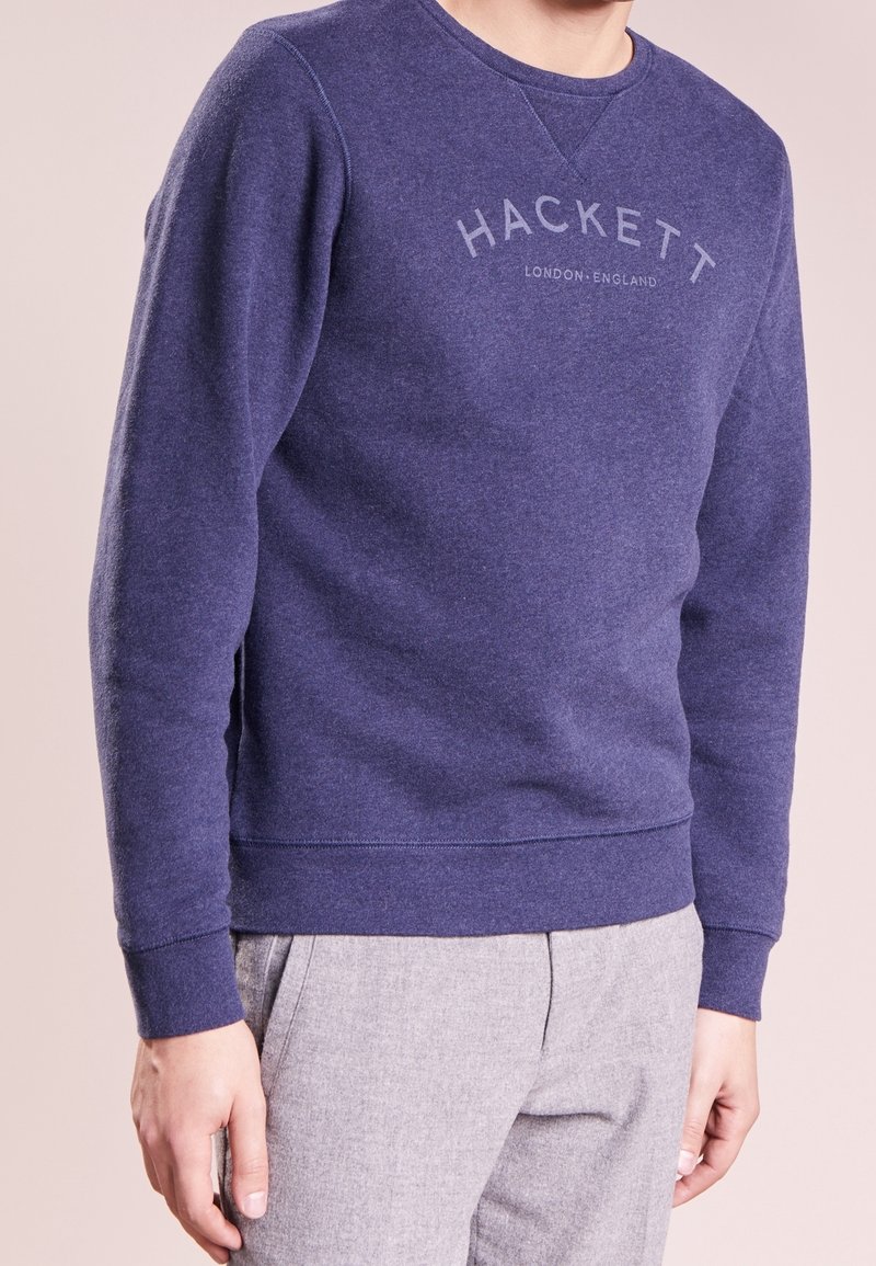 Hombre vestido con un sudadera azul de Hackett London y pantalones gris claro, de pie frente a un fondo neutro.