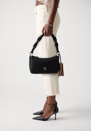 Black textured leather handbag with a twisted handle, silver logo, and a colorful tag. Worn with light-colored cropped jeans and black heels.