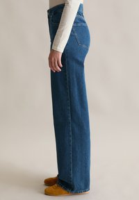 Osoba w wysokich, prostych niebieskich dżinsach, białej bluzce z długim rękawem i musztardowych zamszowych butach stoi bokiem.