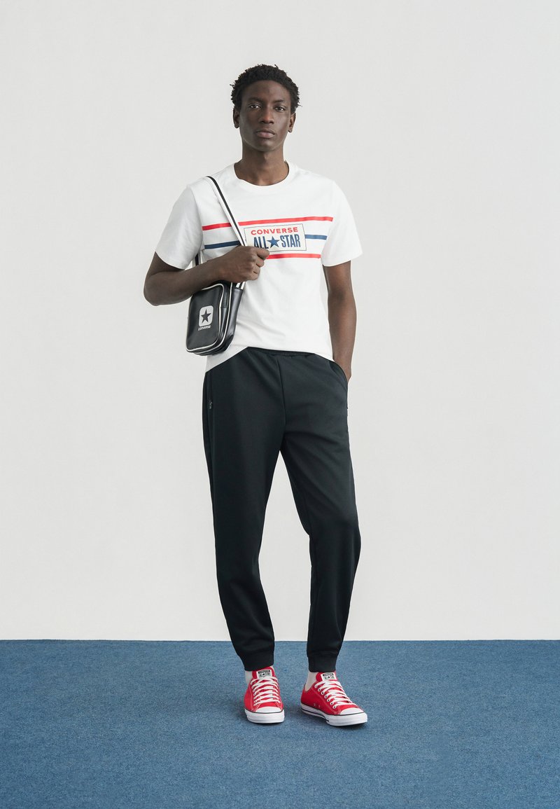 Hvid T-shirt med røde og blå striber og teksten "Converse ALL STAR", sorte træningsbukser, røde sneakers og en sort skuldertaske.