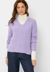 Lavenderfarbener gerippter Strickpullover mit tiefem V-Ausschnitt, langen Ärmeln, die an den Bündchen hochgerollt sind, über einem cremefarbenen Rollkragenpullover getragen, kombiniert mit dunklen Jeans.