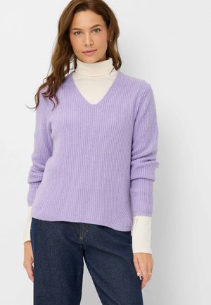 Maglione a coste lavanda con scollo a V profondo, maniche lunghe arrotolate ai polsini, indossato sopra un dolcevita crema, abbinato a jeans scuri.