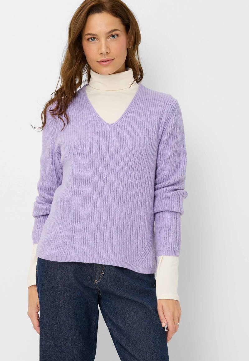 Lavenderfarbener gerippter Strickpullover mit tiefem V-Ausschnitt, langen Ärmeln, die an den Bündchen hochgerollt sind, über einem cremefarbenen Rollkragenpullover getragen, kombiniert mit dunklen Jeans.
