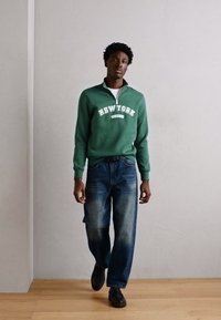 Sweatshirt zippé vert avec le texte blanc "NEW YORK", associé à un jean bleu ample et des chaussures noires, présenté sur un fond neutre.