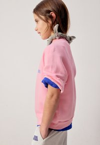 Jeune fille tournée vers la gauche, aux longs cheveux attachés par un nœud gris, portant un sweat-shirt rose aux manches retroussées par-dessus une chemise bleue et un pantalon gris, la main dans la poche.