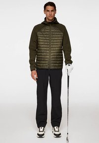 Grön, quiltad jacka med dragkedja framtill, kombinerad med en mörk topp. Svarta byxor och vita skor med svarta detaljer. Golfklubba i handen.