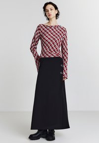 Donna che indossa una camicia a maniche lunghe a quadri rossi, una gonna nera lunga fino alla caviglia con bottoni laterali e stivali neri chunky su uno sfondo semplice.