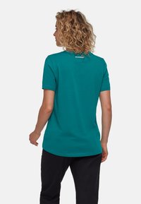 Teal kortärmad t-shirt med rund halsringning och en liten logotyp på baksidan. Enfärgad textur, avslappnad passform, bärs med svarta byxor.