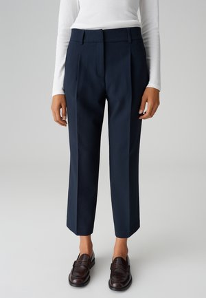 Marineblauwe op maat gemaakte broek met een hoge taille, rechte pijpen en scherpe vouwen. Gemaakt van een gladde stof, gecombineerd met donkere leren instappers.