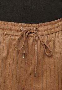 Calças de jogging castanhas com riscas verticais, apresentando uma cintura elástica com um cordão ajustável atado na frente e pontas em metal dourado no cordão.