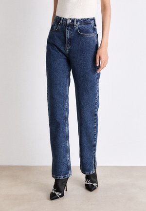 Persoon draagt blauwe spijkerbroek met hoge taille, gecombineerd met zwarte pumps met spitse neus en witte lintdetails.