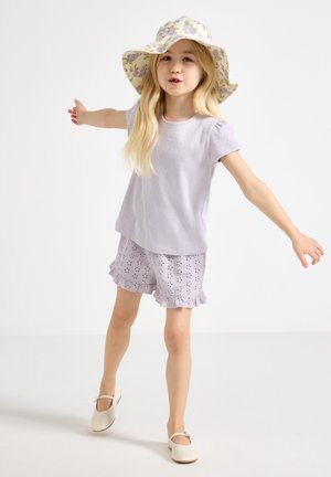 Młoda dziewczynka z długimi blond włosami nosi kwiecisty kapelusz przeciwsłoneczny, lawendowy top z krótkim rękawem, szorty z oczkami i falbankami oraz białe buty Mary Jane, balansując.