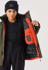 Grön jacka med röd fodring och svarta innermaterial, bärs över en svart topp och svarta byxor med justerbara remmar. Etikett synlig inuti jackan.