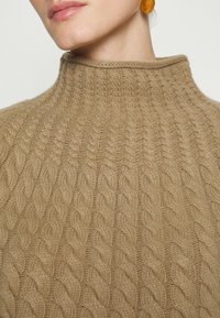 Pull en tricot beige avec un col montant, présentant un motif texturé. Le tissu semble doux et confortable, idéal pour les temps plus froids.