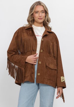 Blonde vrouw draagt een bruine suède fringe jas met knoopsluiting, wit overhemd, lichtblauwe spijkerbroek en gouden oorringen.