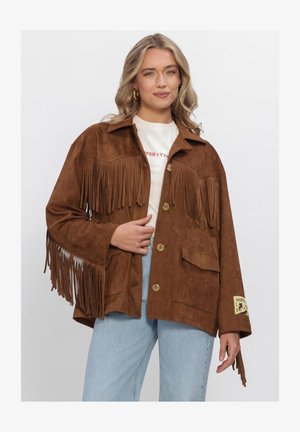 Blonde woman wearing a brown suede fringe jacket with button closure, white shirt, light blue jeans, and gold hoop earrings.