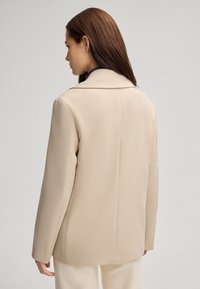 Beige op maat gemaakte blazer met een gestructureerde kraag, lange mouwen en een zachte textuur, met een strakke silhouet en geen zichtbare sluitingen.