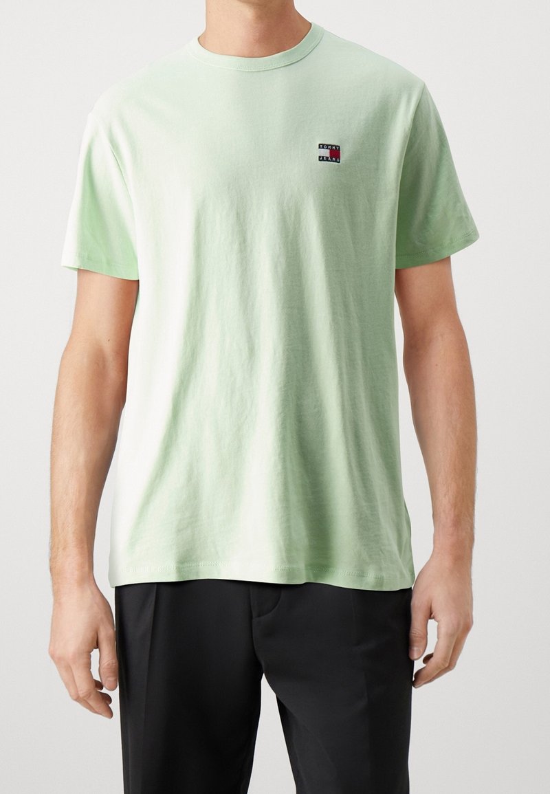 Homme portant un T-shirt vert clair à manches courtes avec un petit logo Tommy Jeans sur la poitrine et un pantalon noir sur un fond uni.