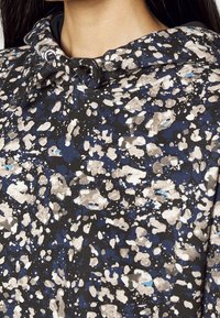 Lähedane pilt inimesest, kes kannab musta, sinist ja beeži laikudega kapuutsi, mille lipsud on seotud kaela ümber rinnaesise sõlme.