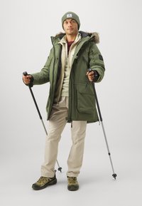 Parka verde con cappuccio foderato in pelliccia, indossato sopra un hoodie chiaro, abbinato a pantaloni beige e scarpe da trekking robuste, che tiene bastoni da trekking.