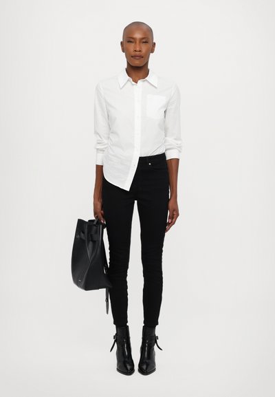 Camisa blanca de botones con un único bolsillo en el pecho, combinada con jeans negros ajustados y botines negros. Lleva un bolso tote negro.