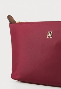 Burgunderfarget nylon-futteral med en gyllen logo-detalj, med en glatt tekstur og enkel, stilren design. Brun skinnkant nær glidelåsen.