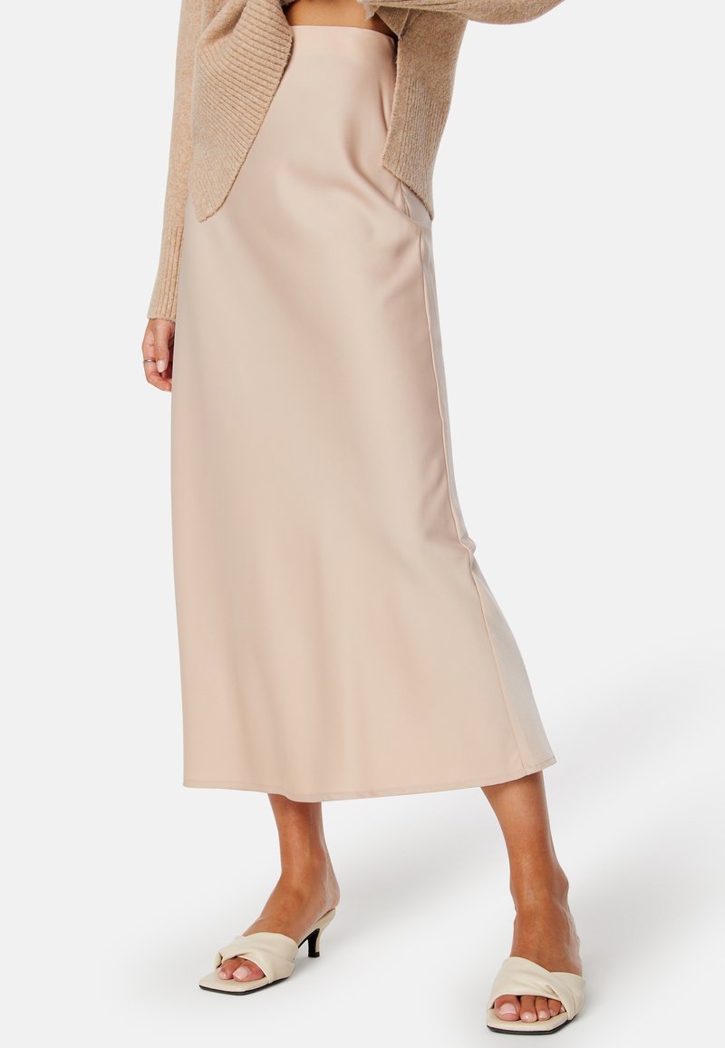 Femme portant une jupe midi beige, un pull en maille beige et des sandales à talons crème sur un fond clair uni.