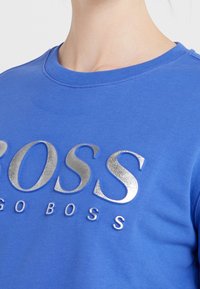 Modrá bavlněná mikina s kulatým výstřihem, zdobená lesklými stříbrnými nápisy "BOSS" a "GO BOSS" ve výrazném, mírně vyvýšeném designu.